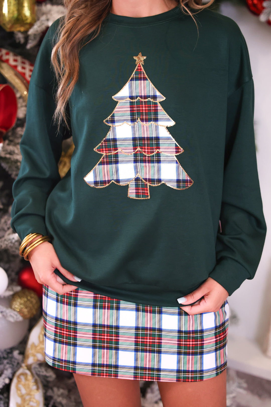 A woman wearing a dark green crewneck sweatshirt with a plaid Christmas tree applique on the front, paired with a matching plaid mini skirt.