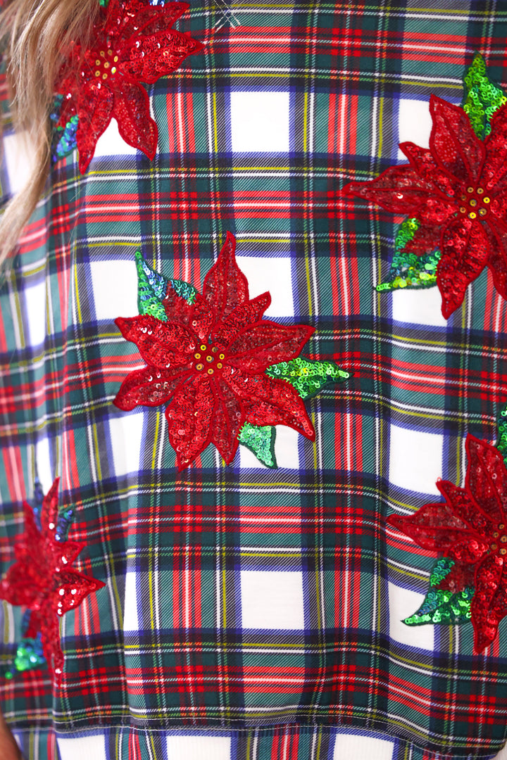 A close-up of a Christmas-themed garment featuring a red, green, and white plaid pattern overlaid with large red sequined poinsettia flowers and green sequin leaves.