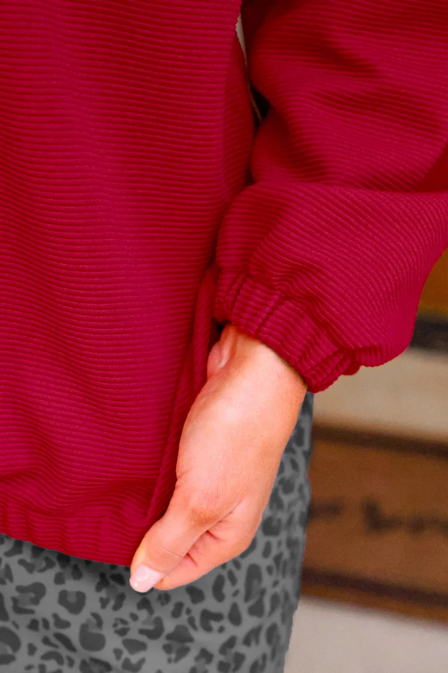 Close-up of a person wearing a bright red ribbed knit top with a cuff sleeve and gray leopard-print bottoms, with one hand tucked into the pocket.