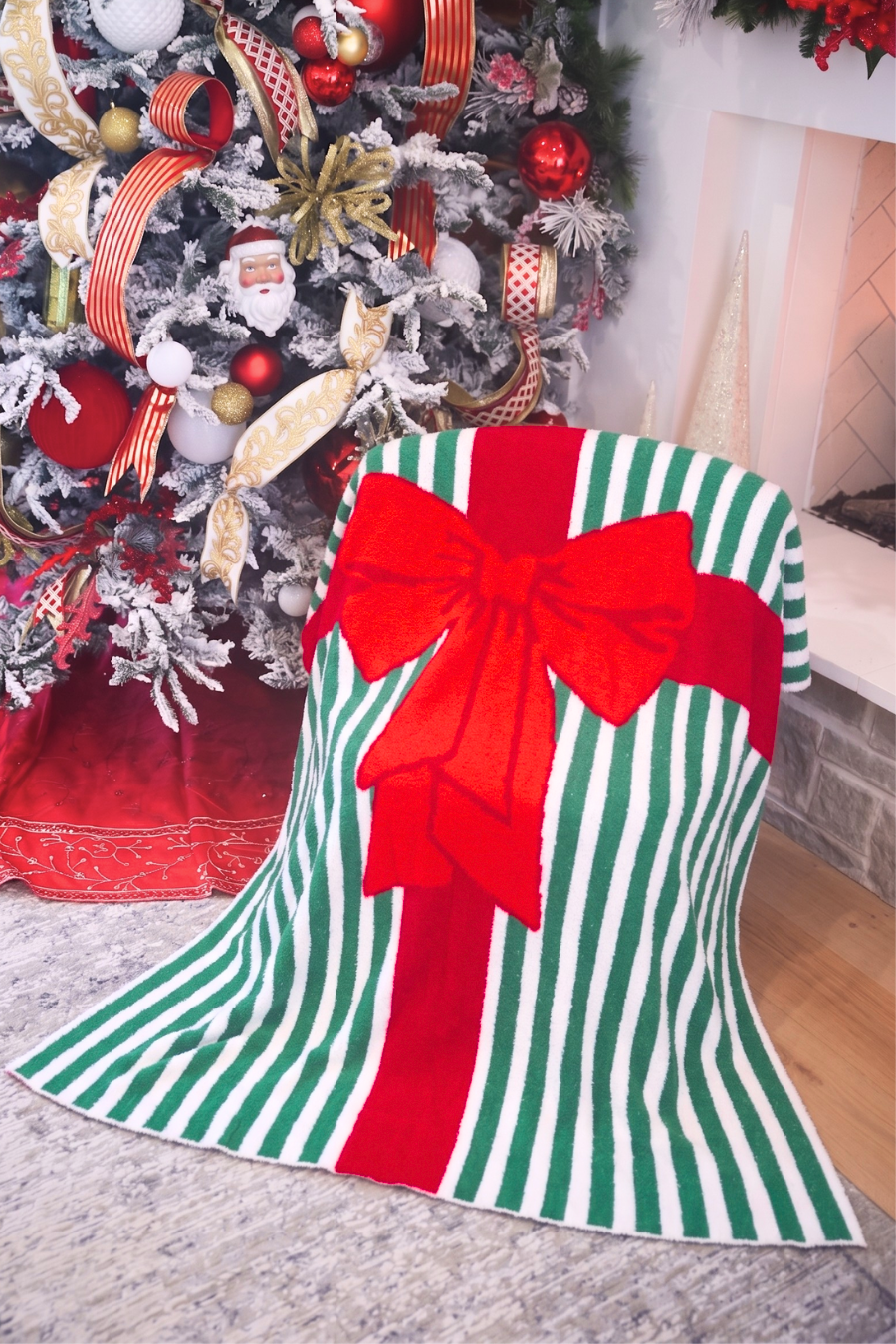 A Christmas-themed bell-shaped throw blanket draped over a chair in a living room setting, featuring a green and white striped pattern with a large red bow design, positioned in front of a decorated Christmas tree.