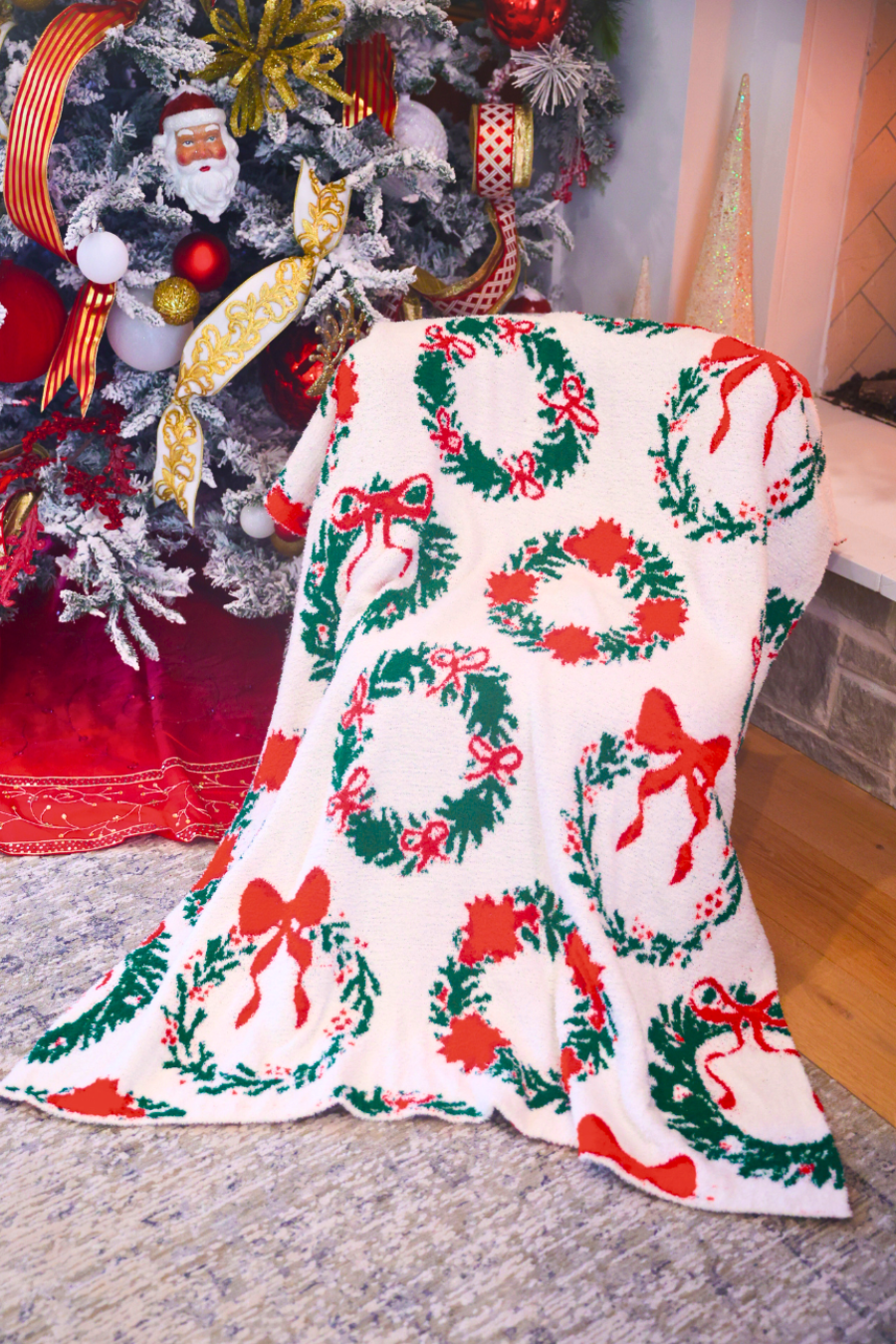 A white fleece throw blanket with a red and green Christmas wreath pattern draped over a chair in front of a decorated Christmas tree.
