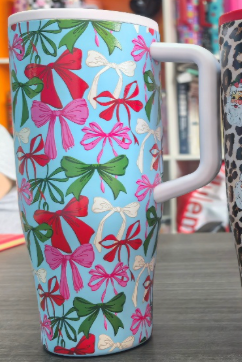 A tall ceramic mug with a white handle, decorated with a repeating pattern of red, pink, and green bows on a light blue background, set on a dark tabletop.
