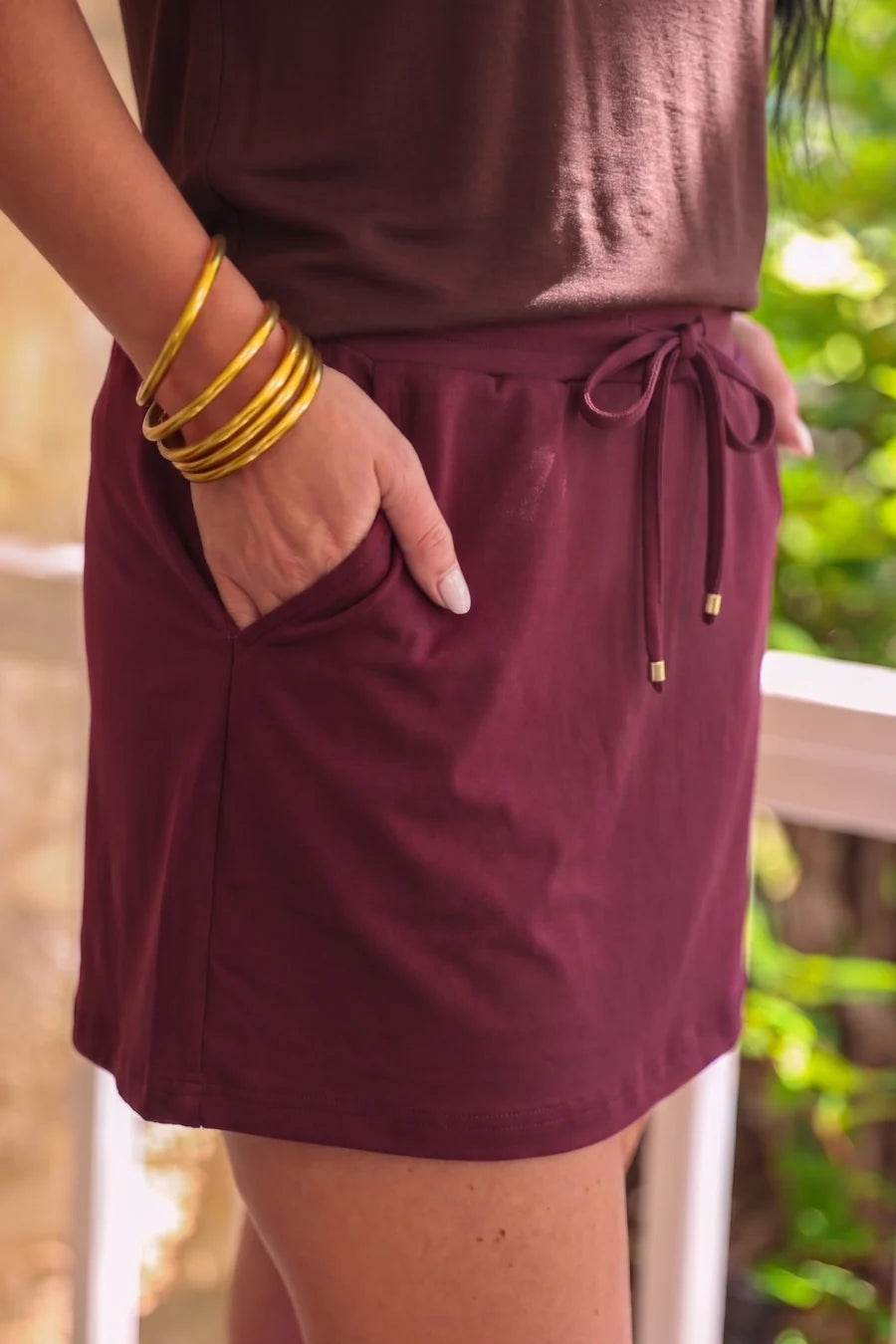Person wearing a burgundy skort with gold bangles in front of a blurred green background