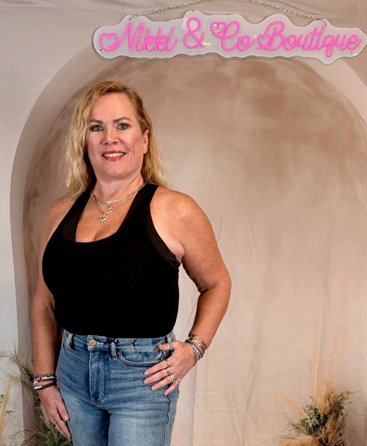 Woman wearing a black tank top and blue jeans standing in front of a beige wall with 'Nikki & Co Boutique' sign.