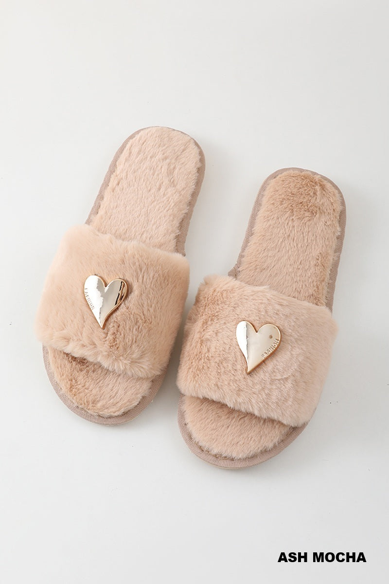 A pair of fluffy open-toe slide slippers in a soft ash-mocha beige color, featuring a metallic gold heart-shaped accent on the strap, shown on a light gray background.