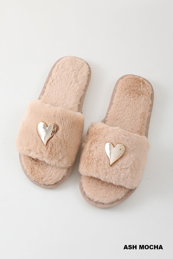 A pair of fluffy open-toe slide slippers in a soft ash-mocha beige color, featuring a metallic gold heart-shaped accent on the strap, shown on a light gray background.