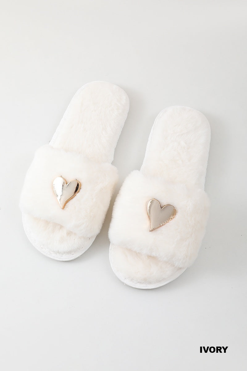 A pair of fluffy ivory slippers with a metallic heart-shaped charm on the top, displayed on a clean white background with the word &