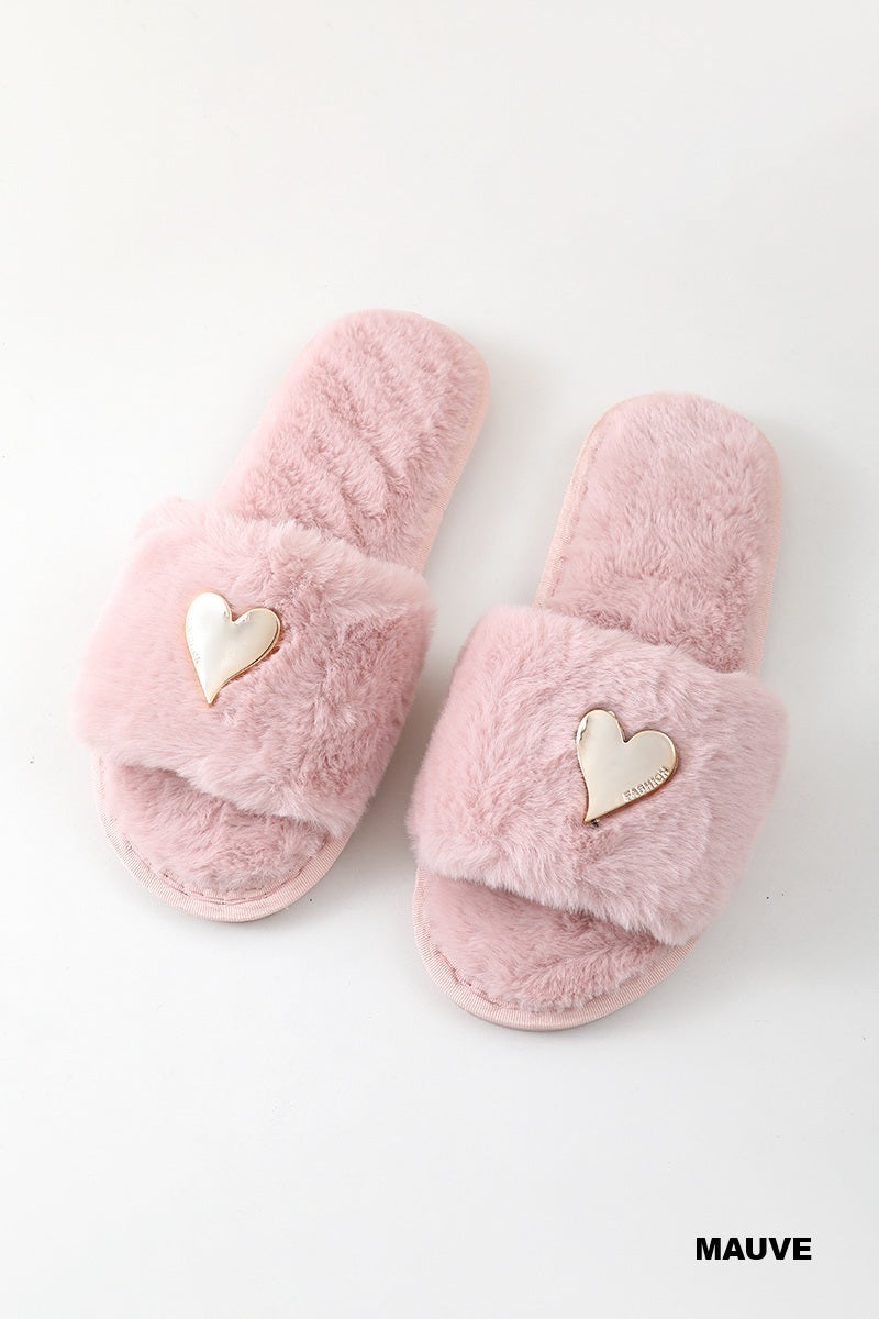 A pair of fuzzy mauve pink slide slippers with a small metallic heart charm on the strap, displayed on a clean white background.