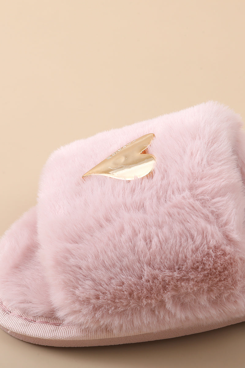 A close-up of a fluffy light pink slippers with a metallic gold heart-shaped accent on the upper, set against a solid beige background.