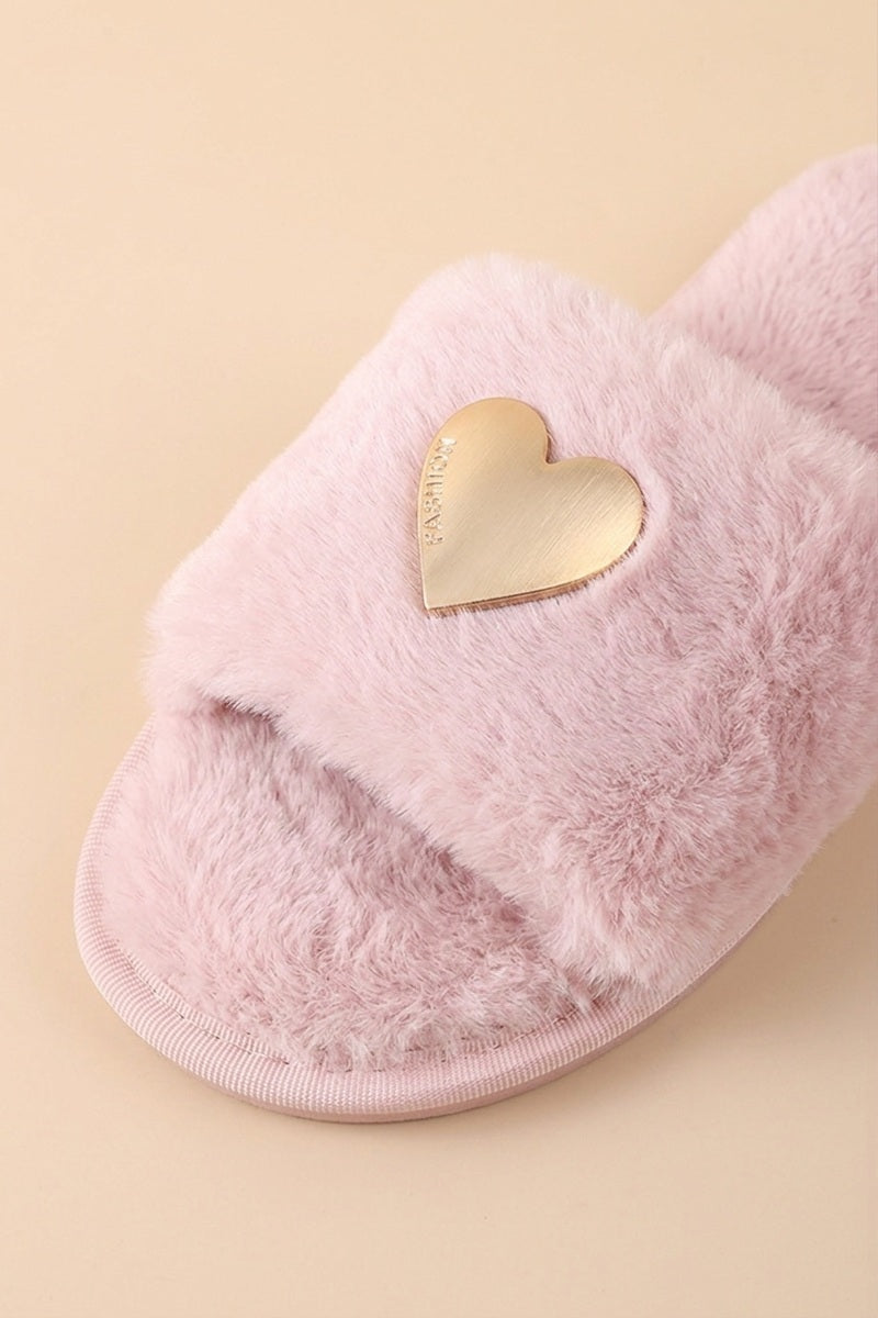 A close-up of a fluffy light pink slide sandal with a metallic gold heart-shaped accent on the strap, set against a solid pale beige background.
