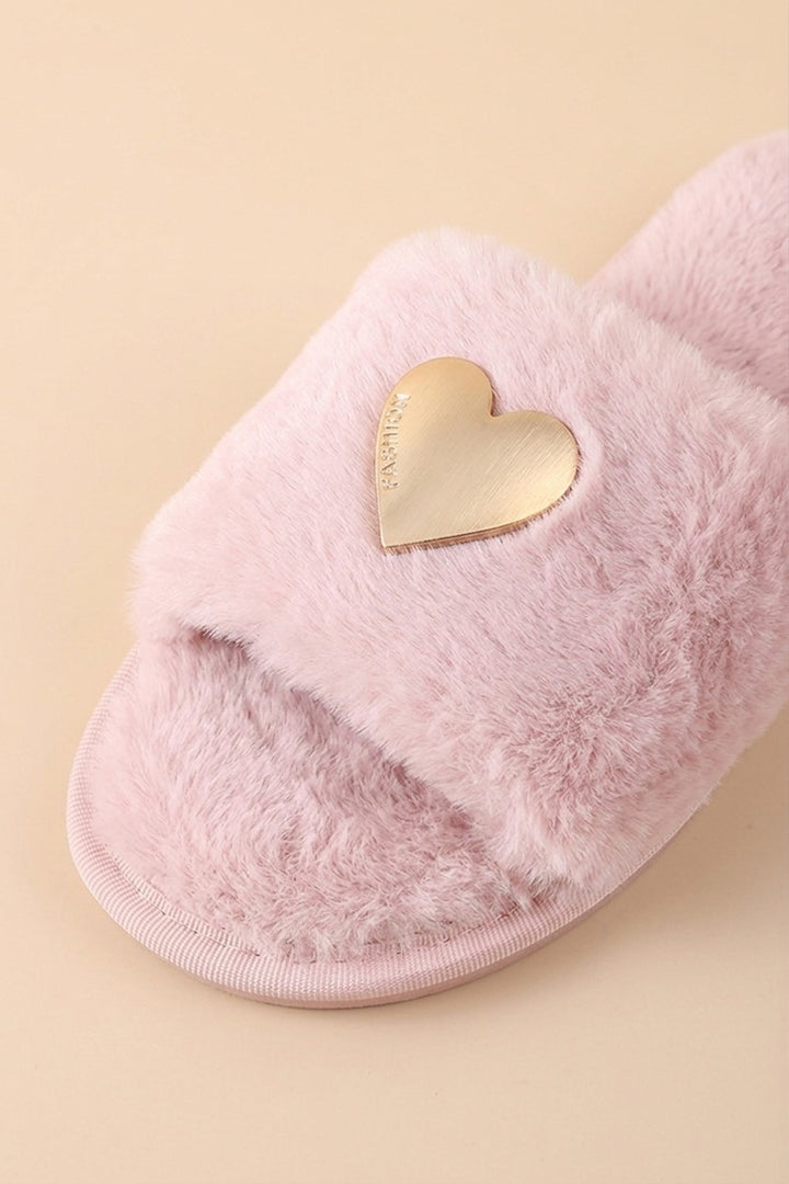 A close-up of a fluffy light pink slide sandal with a metallic gold heart-shaped accent on the strap, set against a solid pale beige background.