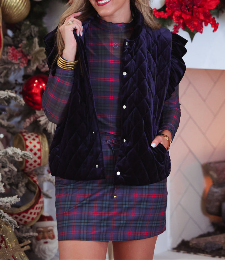 A woman wearing a dark navy velvet quilted vest over a red and blue plaid button-up shirt and matching plaid skort, standing in front of a decorated Christmas tree and fireplace.