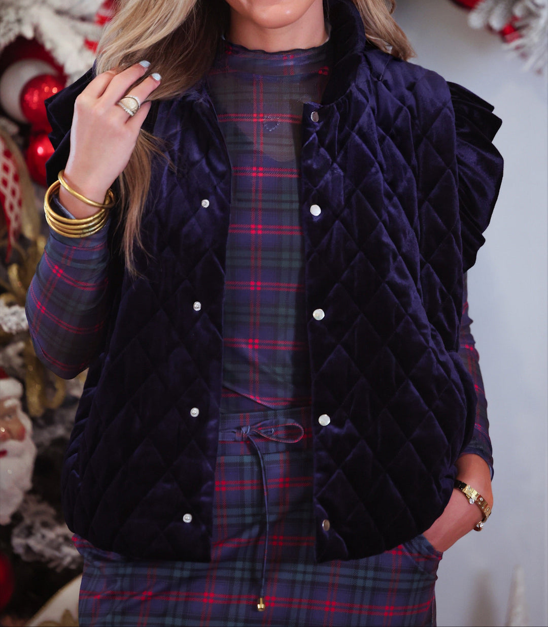 A woman wearing a dark navy quilted velvet vest over a red and green plaid top and matching skort, posed in front of a decorated Christmas tree.