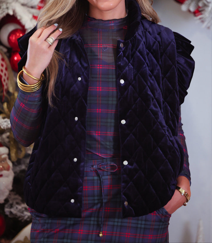 A woman wearing a dark navy quilted velvet vest over a red and green plaid top and matching skort, posed in front of a decorated Christmas tree.