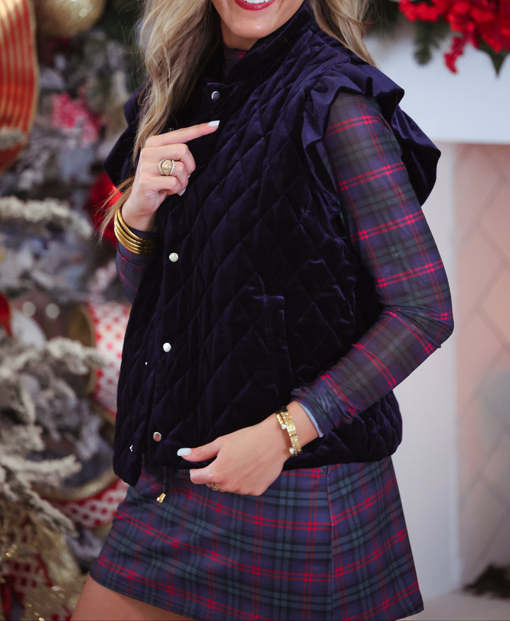 A woman wearing a dark navy quilted sleeveless vest over a red and green plaid long-sleeve shirt and matching plaid skort, standing in front of a decorated Christmas tree and holiday garland.