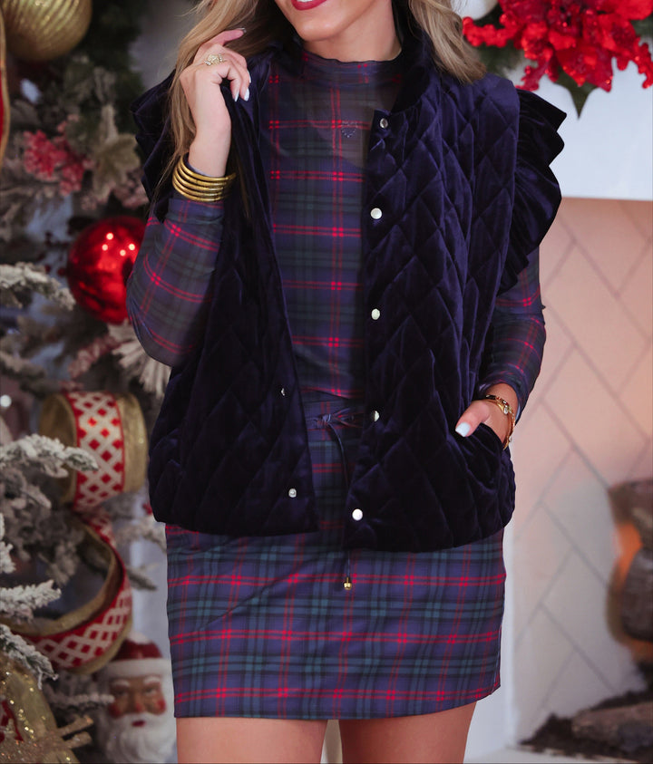 A woman models a dark navy velvet quilted vest over a red and blue plaid long-sleeve top and matching plaid skort, posed in front of a decorated Christmas tree.