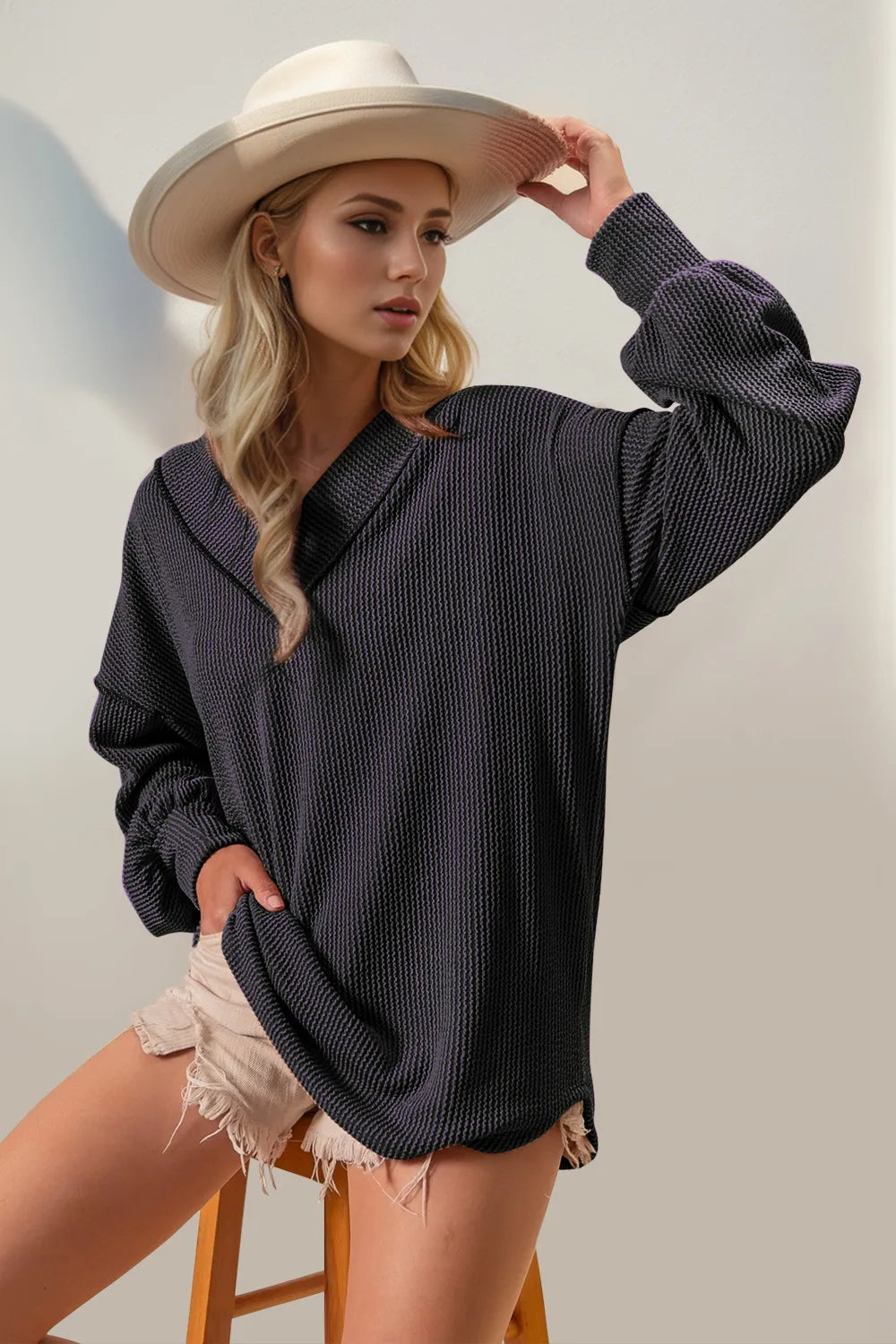 Woman wearing a dark gray oversized sweater and beige hat, standing on a stool against a plain background.