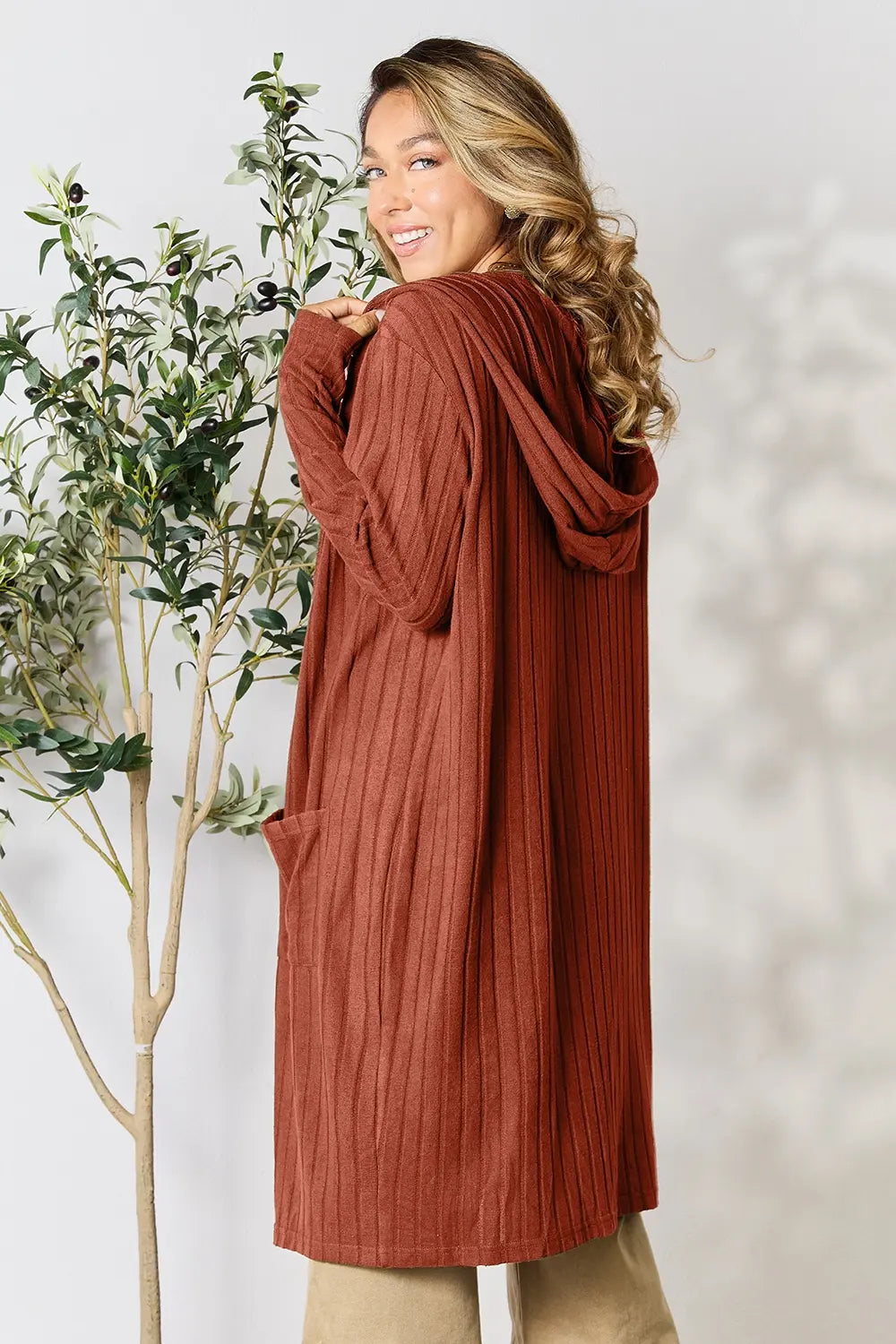 Woman wearing a rust-colored cardigan standing next to a plant against a white background