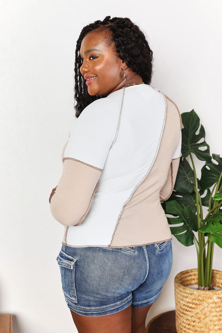 Woman wearing a two-tone shirt and denim shorts standing next to a plant indoors.