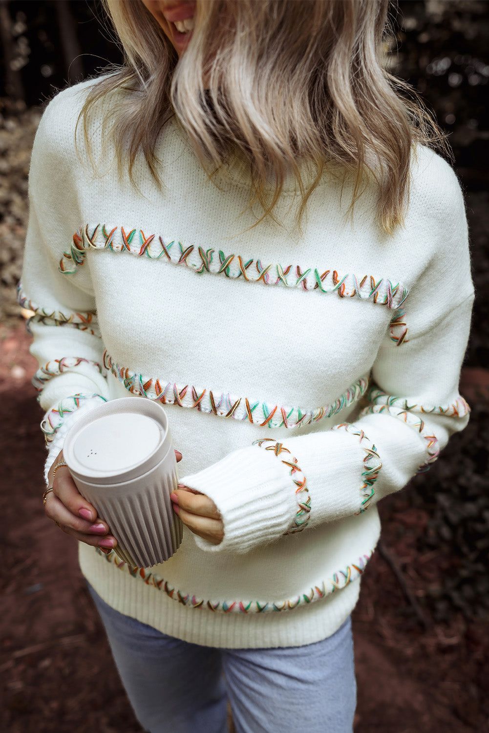 Person wearing a white sweater with decorative patterns, holding a coffee cup outdoors.