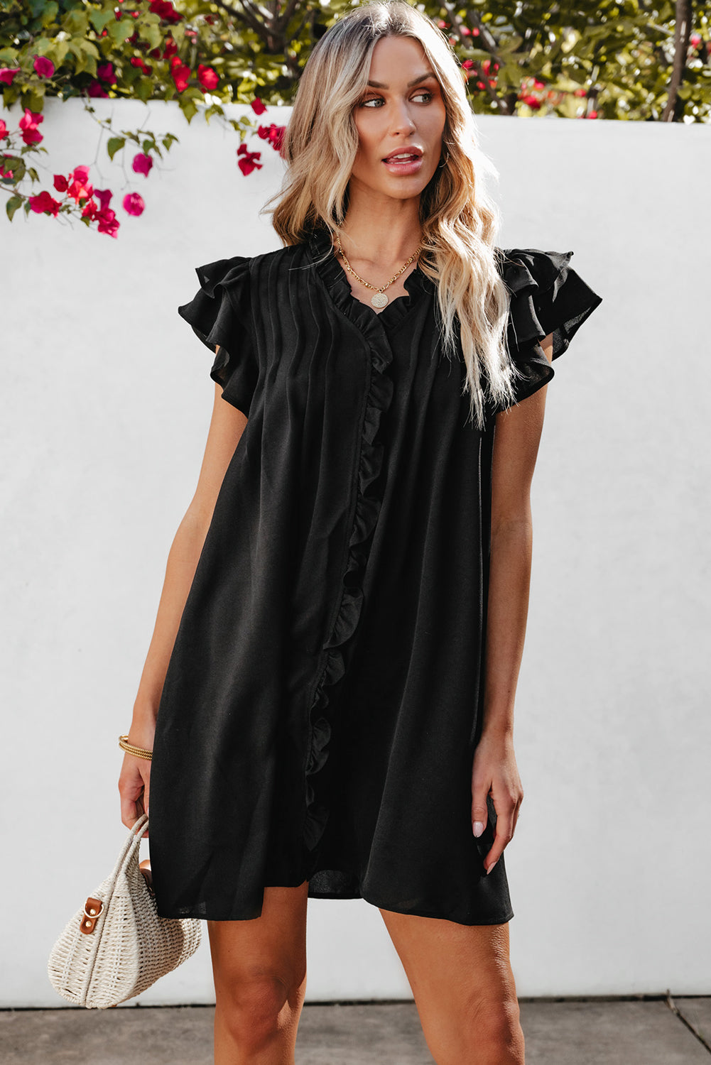 Woman wearing a black dress with ruffled sleeves against a white wall with flowers.