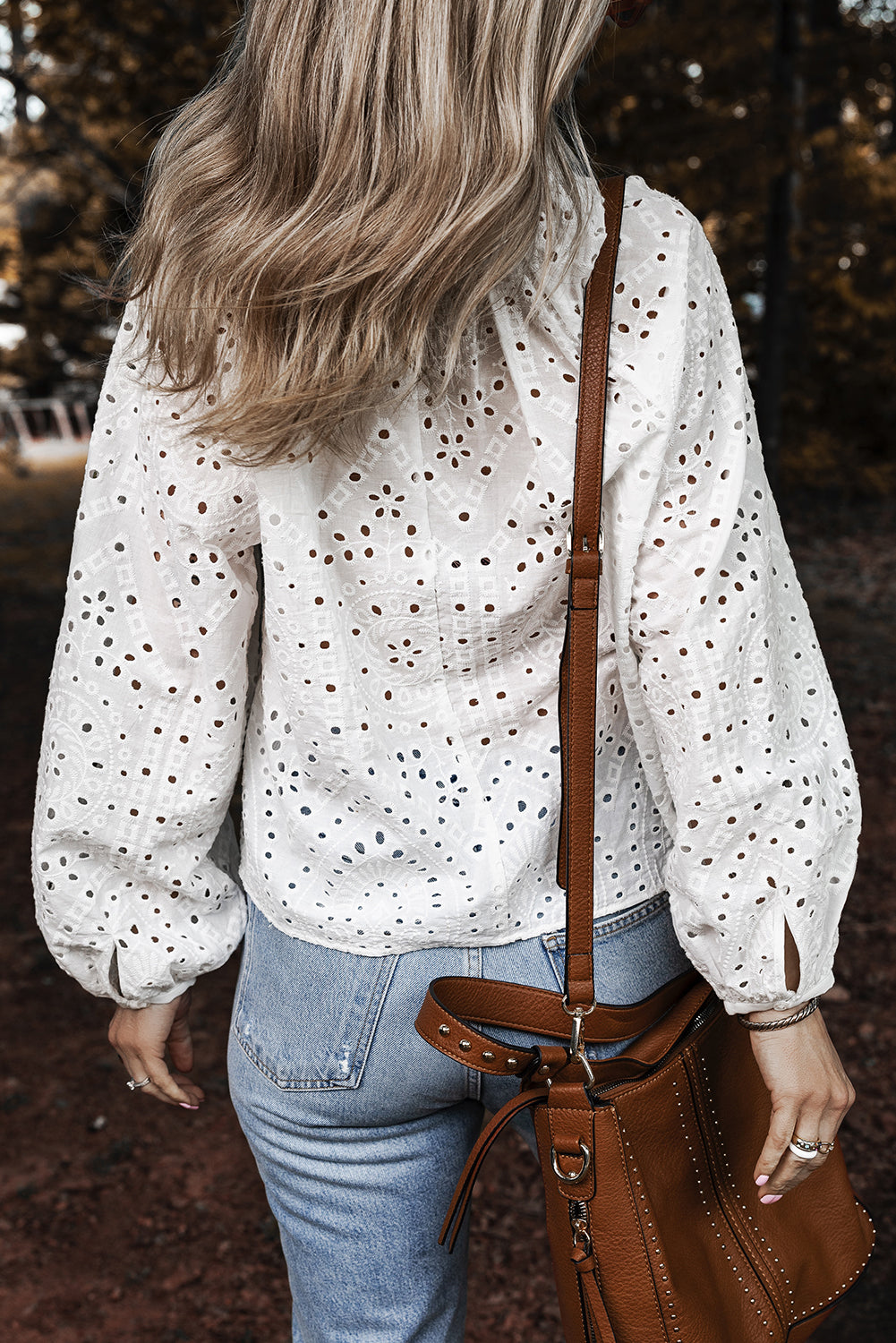 Person wearing a white blouse with eyelet design and blue jeans, holding a brown leather bag.