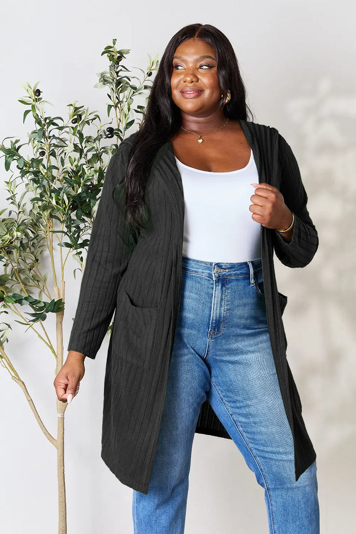 Woman wearing a black cardigan, white top, and blue jeans standing next to a plant.
