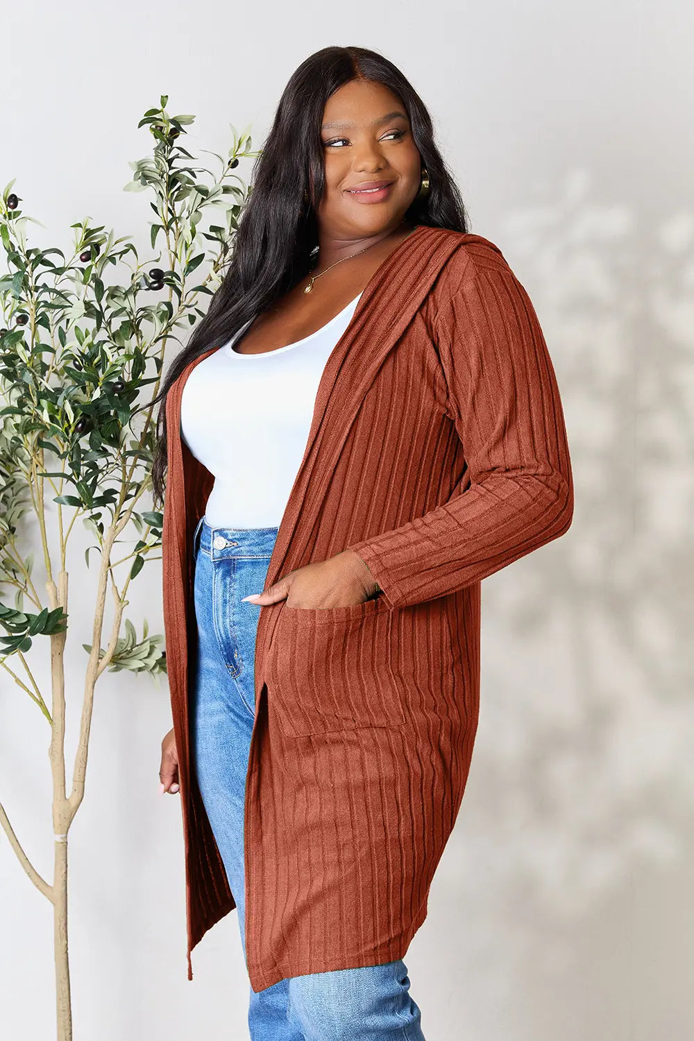 Woman wearing a rust ribbed cardigan over a white top and blue jeans, standing against a neutral background with a plant.