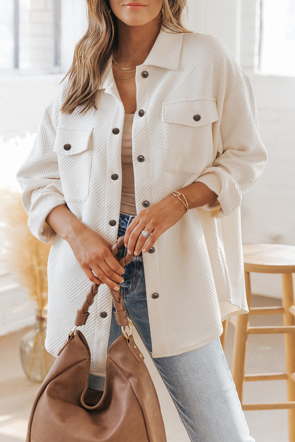 Woman wearing a beige textured jacket with black buttons, holding a brown handbag indoors.