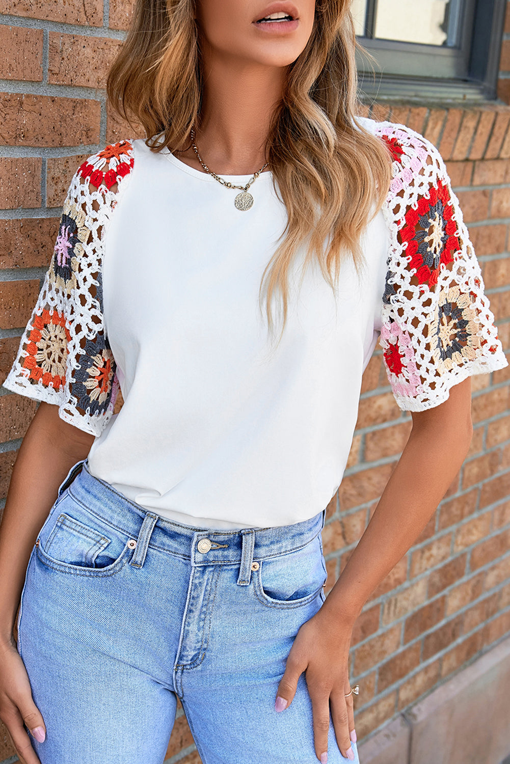 Woman wearing a white top with colorful crochet sleeves and blue jeans against a brick wall.
