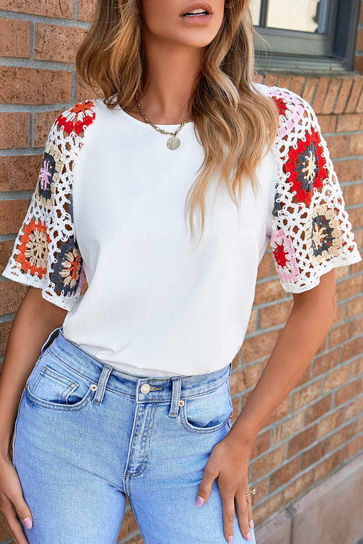 Woman wearing a white top with colorful crochet sleeves and blue jeans against a brick wall.