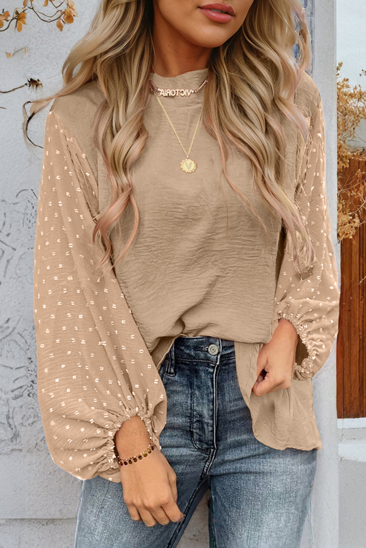 Woman wearing a beige long-sleeve top with polka dot pattern, beige shirt, and blue jeans against a light gray wall.
