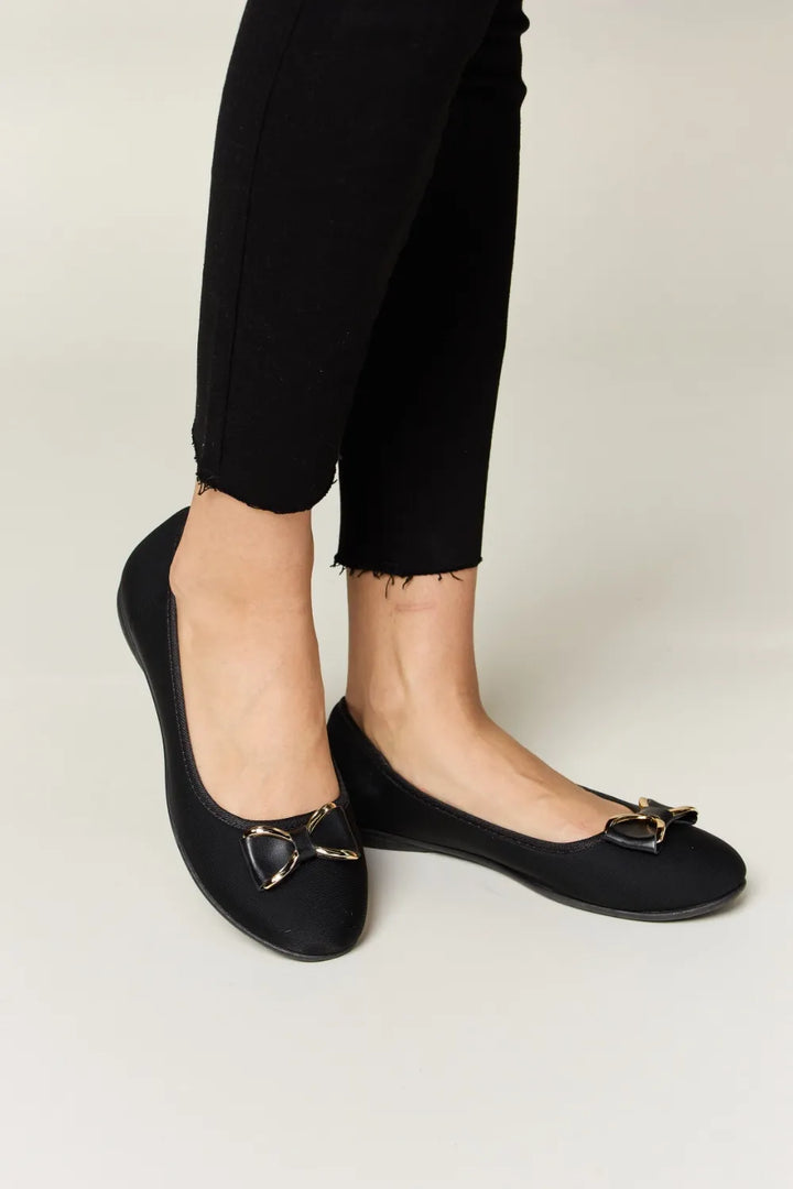 Black flat shoes with decorative buckles worn with black pants on a light background