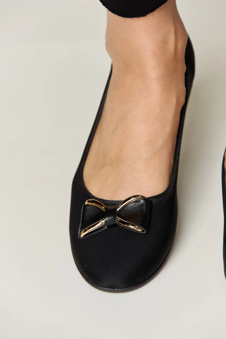 Black flat shoes with gold bow detail on a light gray background