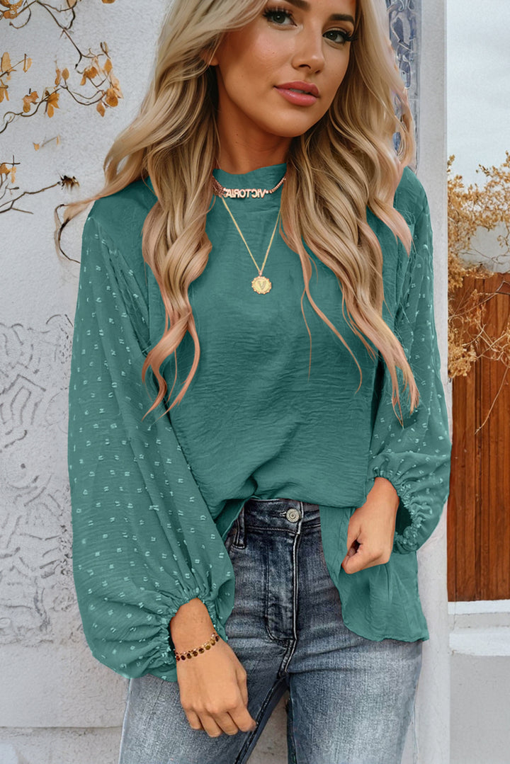 Woman wearing a teal long-sleeve top with a subtle pattern, standing against a decorative wall.