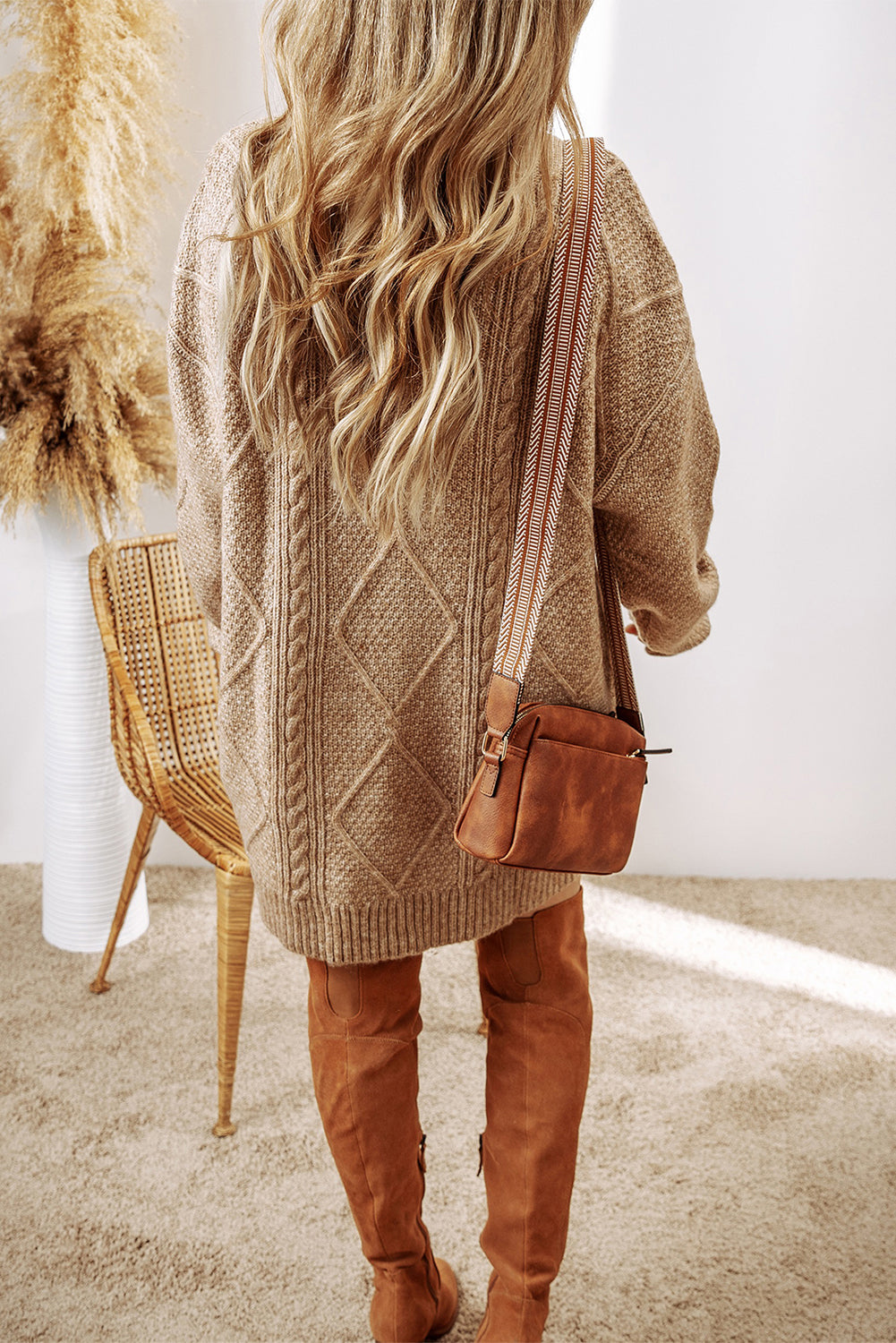 Person wearing a beige cardigan dress and brown boots, holding a brown handbag indoors.