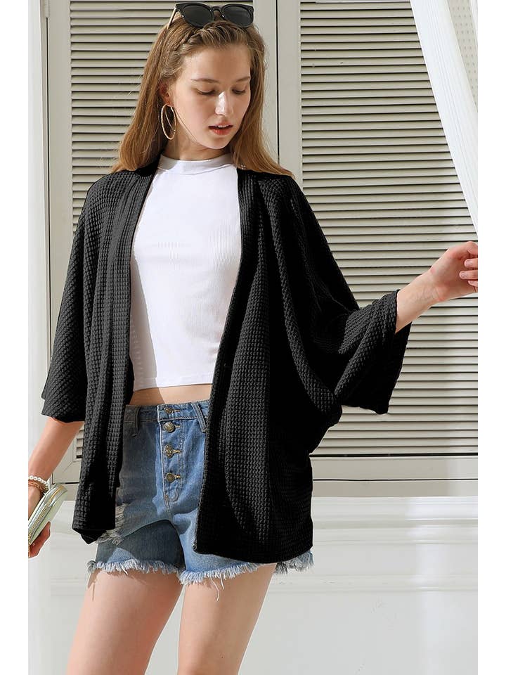 Woman wearing a black cardigan, white top, and denim shorts outdoors.