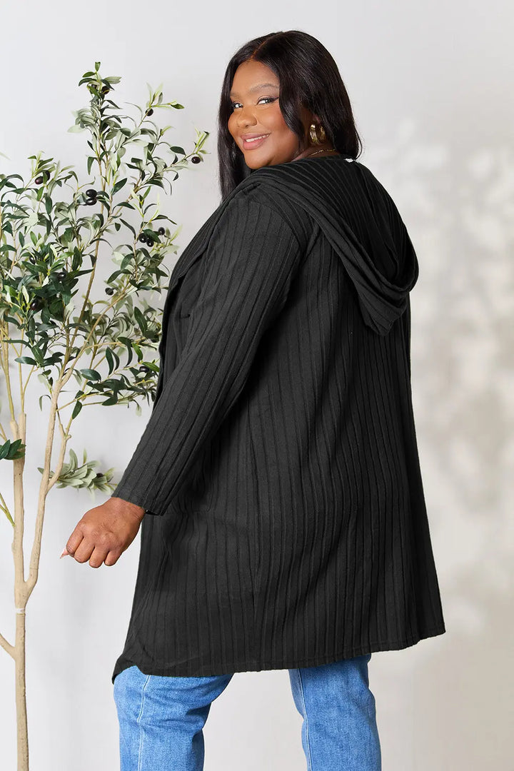 Woman wearing a black ribbed cardigan over a white top and blue jeans, standing next to a plant.