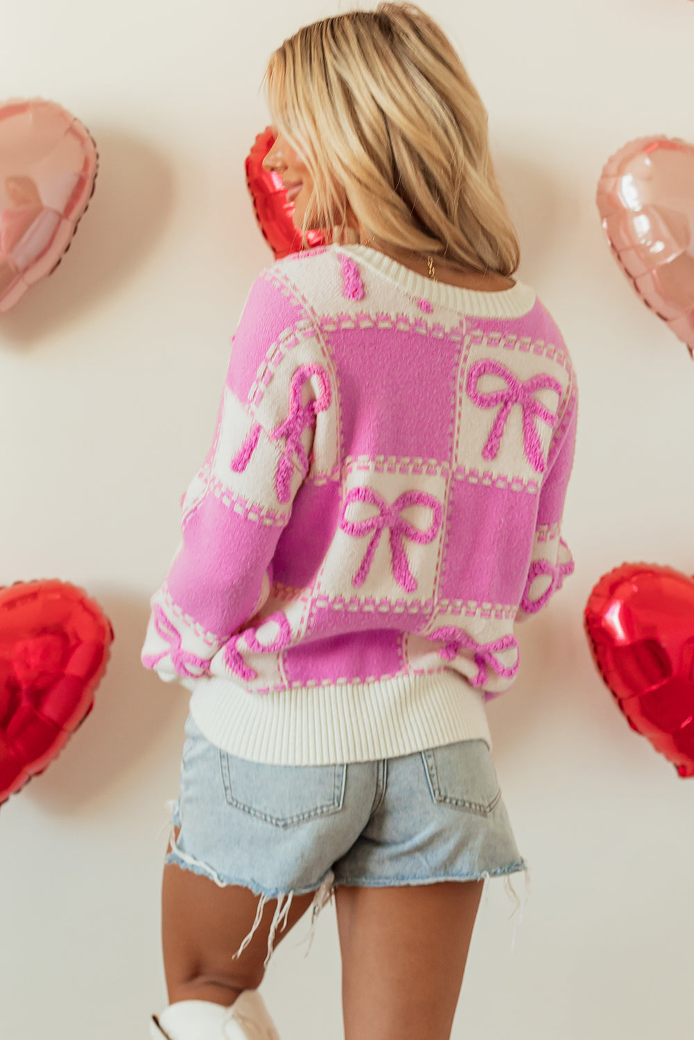 Person wearing a pink and white sweater with bow patterns, standing against a light background with heart-shaped balloons.