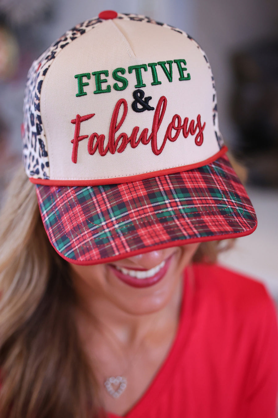 A woman wearing a festive trucker hat with a cream front panel, leopard print side panels, a red plaid visor, and embroidered text reading &