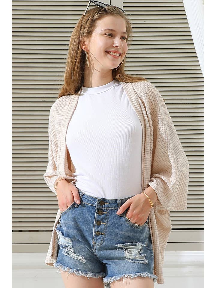 Woman wearing a coconut cardigan, white shirt, and blue shorts against a neutral background