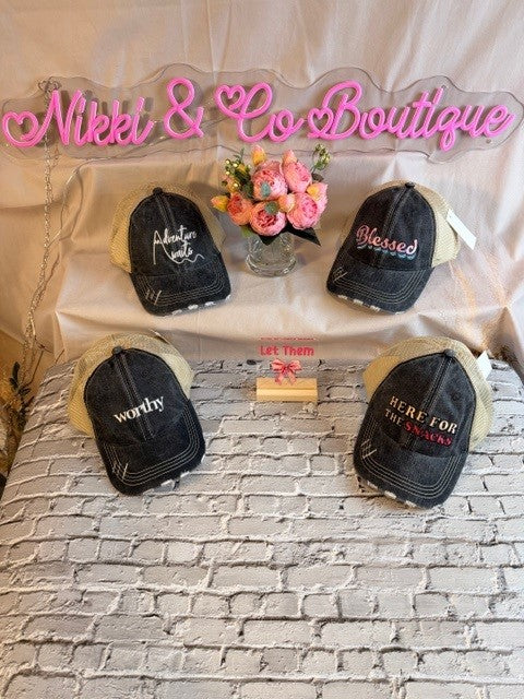 Black baseball caps with embroidered text on a white brick surface with 'Nikki & Co Boutique' sign.