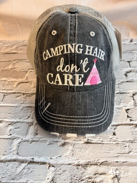 Black cap with 'Camping Hair Don't Care' text and pink teepee graphic on a stone wall background