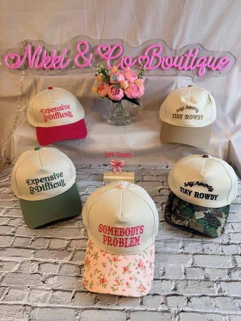 Five baseball caps with different text designs on a surface with 'Nikki & Co Boutique' sign in the background.