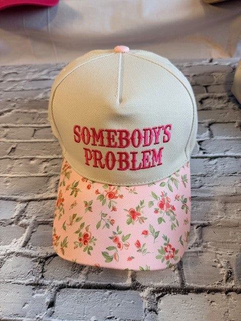 Cap with 'Somebody's Problem' text on floral pink fabric against a brick wall.
