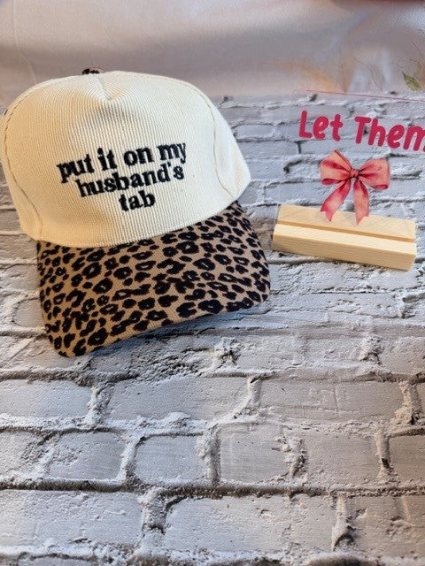 Cap with leopard print brim and text 'Put it on my husband's tab' on a textured surface.