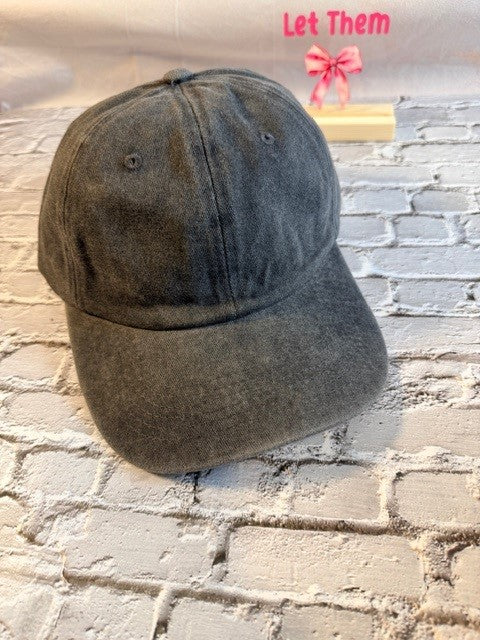 Dark gray baseball cap on a textured stone surface with 'Let Them' text and pink bow in the corner.