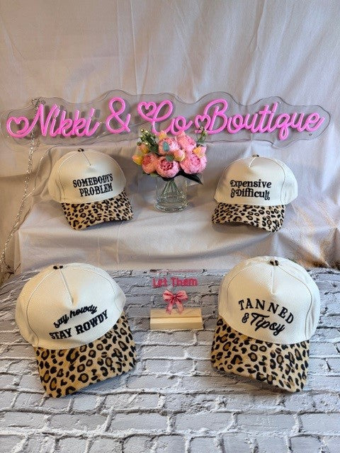 Four leopard print baseball caps with text on a white and gray brick background with 'Nikki & Co Boutique' sign.