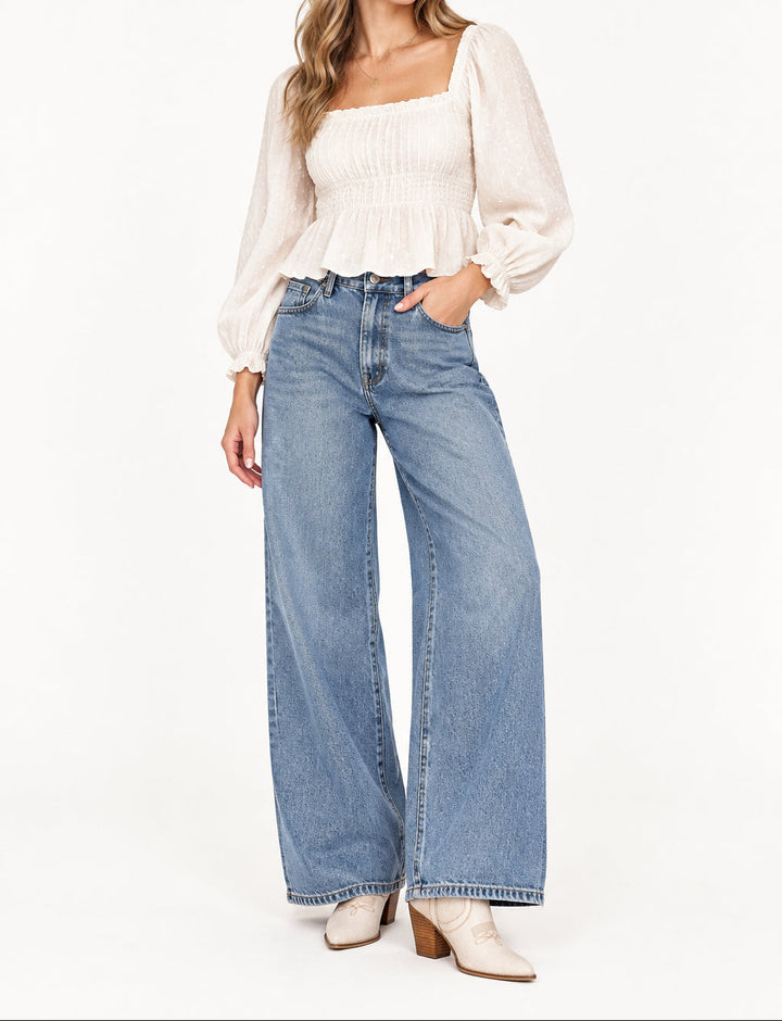 A person wearing light beige western-style ankle boots with pointed toes, decorative stitching, and stacked brown block heels, paired with cuffed light blue jeans against a white background.